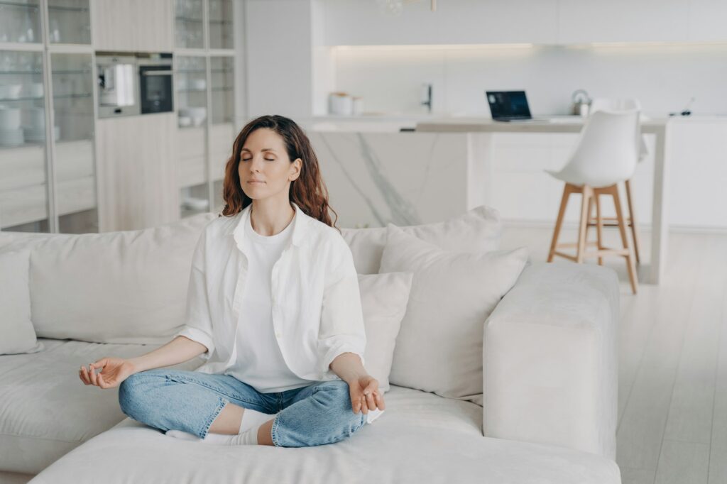 Hispanic woman practicing yoga, meditates, doing breathing exercises on comfortable sofa at home